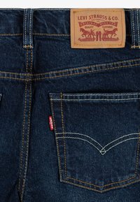 Tamno plavi traperice s tamnom kožnom etiketom na kojoj stoji tekst "LEVI STRAUSS & CO." i žutim šavovima. Stražnji džepovi imaju zakrivljeni dizajn.