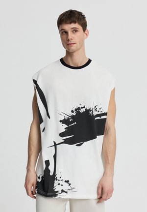 Giovane uomo che indossa una maglietta bianca senza maniche con un motivo astratto di schizzi di vernice nera, in piedi davanti a uno sfondo chiaro semplice.