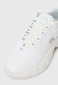 Białe siatkowe buty sportowe z teksturowanej tkaniny, subtelnymi szarymi akcentami oraz logo po boku. Podeszwa jest gruba, co zapewnia amortyzację.
