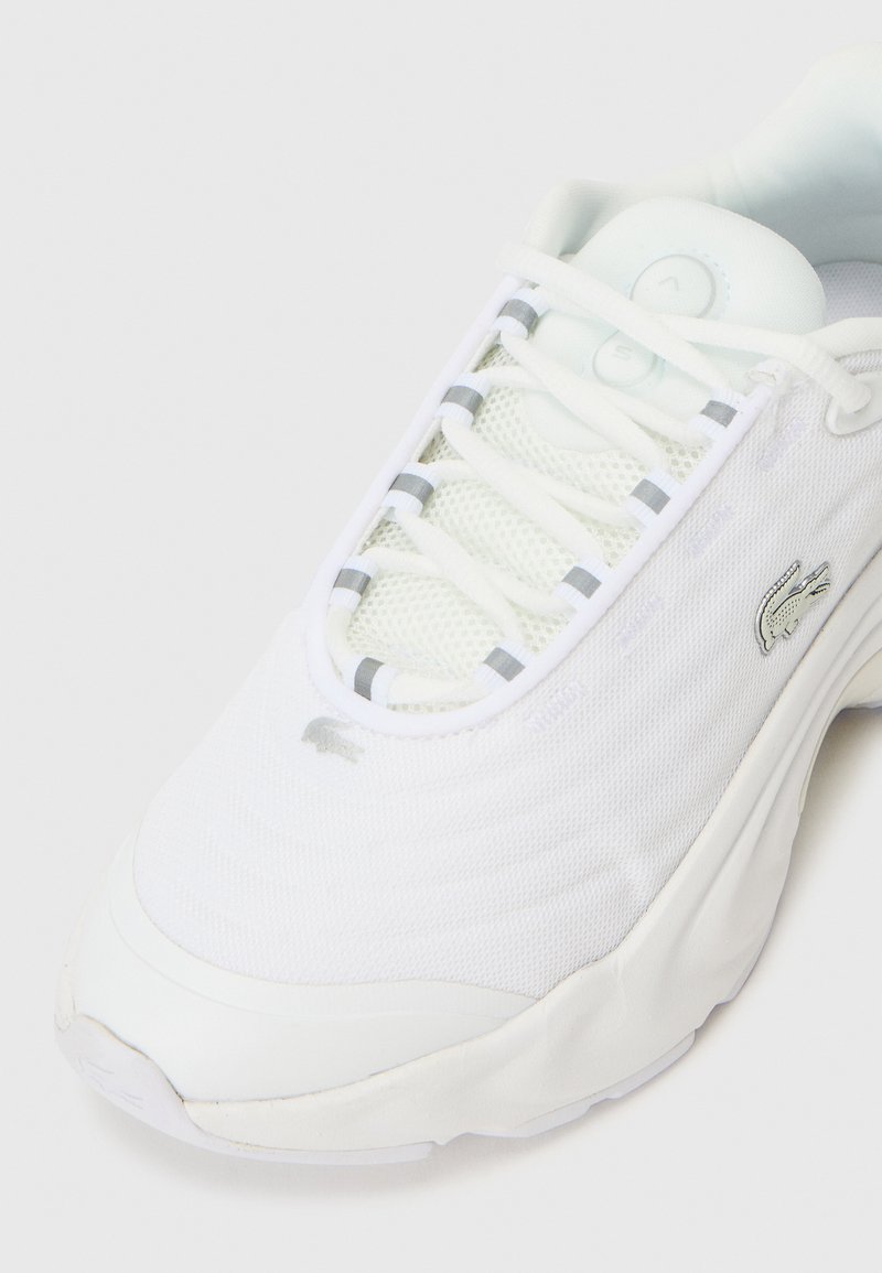 Zapatilla de malla blanca con tejido texturizado, sutiles acentos grises y un logo en el lateral. La suela es gruesa, lo que proporciona amortiguación.
