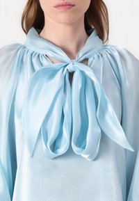 Blusa di raso azzurro chiaro con scollatura arricciata e grande dettaglio a fiocco sul davanti. Tessuto liscio con una finitura lucida.