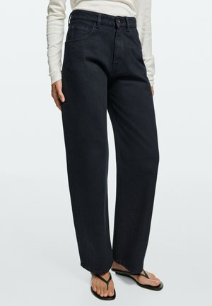 Donna che indossa jeans neri a vita alta a gamba larga, top bianco a maniche lunghe e sandali neri su sfondo semplice.