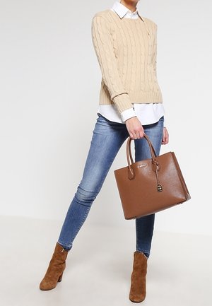 Kvinde iført beige kabelstrikket sweater, hvid skjorte, blå skinny jeans, brune ruskindsankelstøvler, holder stor brun læderhåndtaske.