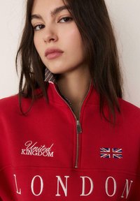 Felpa rossa con zip, con ricami bianchi con scritte "LONDON" e "United Kingdom", e piccolo dettaglio della bandiera Union Jack sul petto.