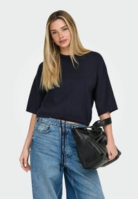 Pull court bleu marine à manches courtes avec ourlet à cordon de serrage, associé à un jean large bleu clair. Un sac à main en cuir noir est tenu à la main.