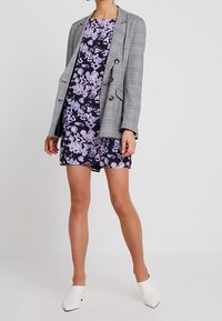 Model w szarym kraciastym żakiecie na kwiecistej granatowej sukience, w białych mule na stopach, pozująca na jednolitym tle.