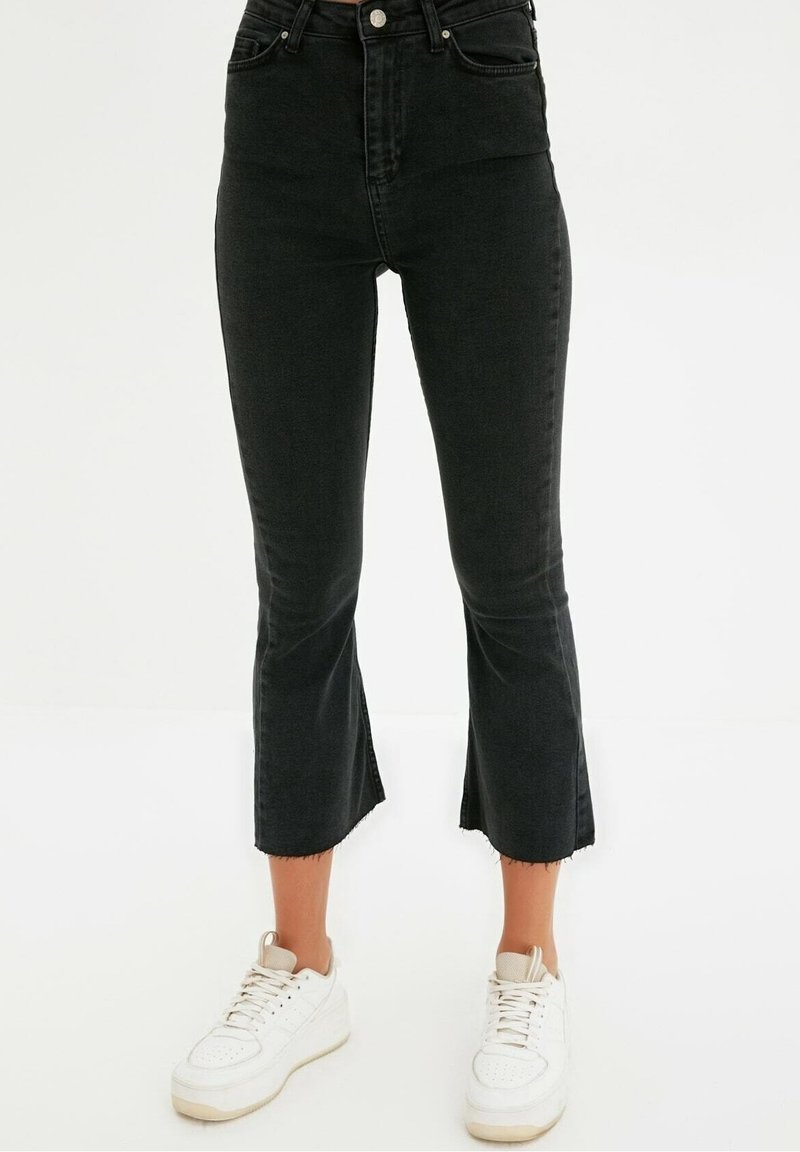 Vaqueros negros de tiro alto con un dobladillo acampanado, hechos de denim, con un borde sin rematar en la parte inferior, combinados con zapatillas deportivas blancas.