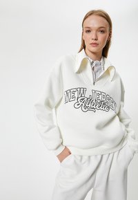 Biała, oversize'owa bluza z półzamkiem i wysokim kołnierzem. Posiada czarny napis "NEW JERSEY Athletic". Połączona z pasującymi białymi spodniami.
