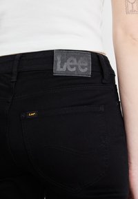 Zbliżenie na czarną tylną kieszeń spodni Lee i metkę na pasku u osoby noszącej biały top, z widoczną częścią ramienia i tatuażem.