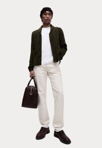 Grün gestrickte Strickjacke mit Taschen, weißes T-Shirt, cremefarbene Hose, braune Lederschuhe und eine dunkelbraune Handtasche mit goldfarbener Akzentuierung.