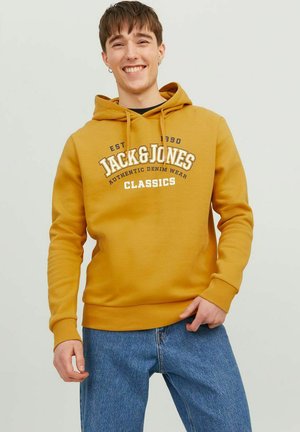Gelbes Kapuzensweatshirt mit Kängurutasche, das einen weißen und schwarzen Logo-Text trägt. Getragen mit blauen Jeans. Glatte Stoffstruktur.
