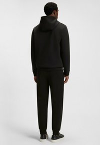 Uomo di spalle che indossa una felpa con cappuccio nera, pantaloni joggers neri abbinati e sneakers nere con suole bianche su uno sfondo neutro.