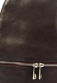 Le sac en cuir marron présente une surface texturée, une fermeture éclair en argent et deux tirettes de fermeture éclair. Design compact et minimal avec des lignes épurées.