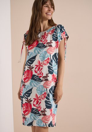 Donna sorridente che indossa un vestito corto con maniche a giro fino al ginocchio con una stampa colorata di foglie e fiori tropicali su uno sfondo chiaro.