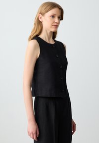 Gilet in lino nero senza maniche con scollo rotondo e cinque bottoni frontali. Progettato con una silhouette corta e una texture liscia.