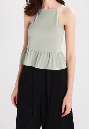 Vrouw draagt een lichtgroene mouwloze peplum top gecombineerd met zwarte, high-waisted, wijde broekspijpen tegen een effen achtergrond.