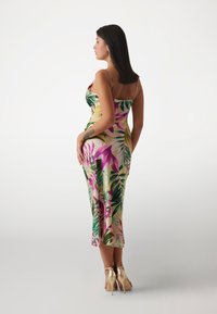 Vestido de satén con un patrón floral tropical en verde, rosa y amarillo. Silueta ajustada con tirantes finos. Usado con zapatos de tacón alto dorados.