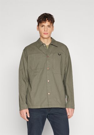 Olivgrüne Button-Up-Jacke aus leichtem Material, mit zwei Taschen auf der Vorderseite, Kupferknöpfen und einem kleinen Logo auf der Brust.