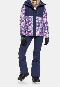 Bunte Schneejacke mit lila und rosa Blumenmustern, kombiniert mit navyblauen Skihosen. Schwarze Handschuhe und Winterstiefel runden das Outfit ab.
