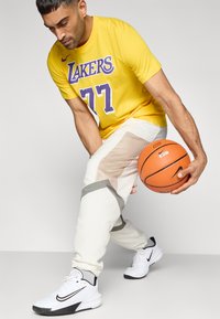 Κίτρινη μπλούζα με την επιγραφή "Lakers" και τον αριθμό 77 σε μωβ, συνδυασμένη με λευκό παντελόνι με διχτυωτό ύφασμα και γκρι λεπτομέρειες, κρατώντας μια μπάλα του μπάσκετ.