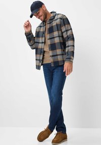 Geruite shirtjas in navy, grijs en tan; tan gebreide trui; blauwe spijkerbroek; bruine suède schoenen; navy pet; casual houding met hand omhoog.