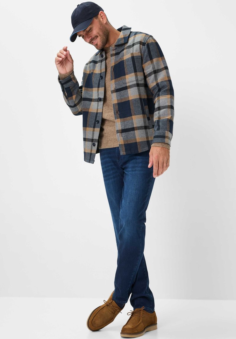 Geruite shirtjas in navy, grijs en tan; tan gebreide trui; blauwe spijkerbroek; bruine suède schoenen; navy pet; casual houding met hand omhoog.