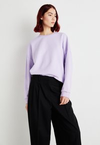 Lavanda oversized dukserica s spuštenim ramenima, rebrastim manžetama i okruglim izrezom, u kombinaciji s širokim crnim hlačama. Jednostavan i moderan dizajn.
