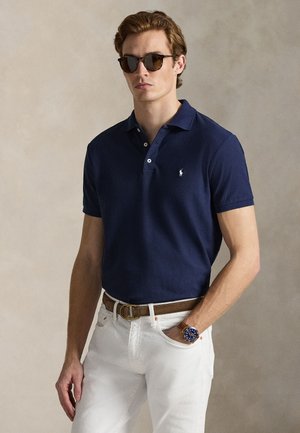 Άνδρας που φοράει μπλε μπλούζα πόλο, λευκό παντελόνι με καφέ ζώνη, γυαλιά ηλίου και ρολόι χειρός, στέκεται μπροστά σε απλό μπεζ φόντο.