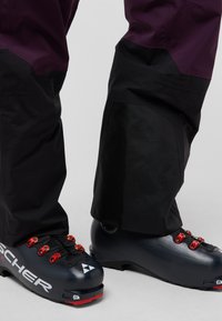 Gli scarponi da sci presentano un'esterno nero e grigio scuro con hardware rosso, pannelli testurizzati e un logo distintivo. I pantaloni sono neri con un accento viola.