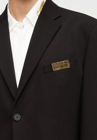 Μαύρο σακάκι με χρυσή πλακέτα «Versace Jeans Couture» στην τσέπη του στήθους, φορεμένο πάνω από λευκό πουκάμισο με διακοσμητικό γιακά.