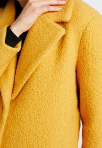 Cappotto giallo in misto lana con una superficie texturizzata e morbida. Presenta un colletto a revers e un'apertura frontale, parzialmente oscurata da una mano.