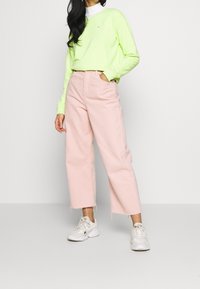 Maglietta a maniche lunghe verde neon con un piccolo logo, abbinata a pantaloni rosa chiaro a gamba larga e scarpe da ginnastica bianche. Outfit casual con colori a contrasto.