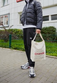 Osoba nosząca czarną kurtkę i spodnie North Face, białe trampki, trzyma beżową torbę na ramię z powtarzającym się słowem "CHANGE" w czarnym i czerwonym tekście.