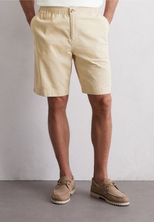 Mand iført beige elastiske shorts med knap foran og lommer, kombineret med brune snørestøvler på gråt gulv.