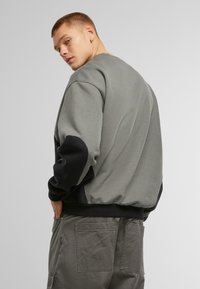 Sudadera gris con mangas negras acentuadas, puños y dobladillo acanalados, y un ajuste relajado. El material parece suave, diseñado para uso casual.