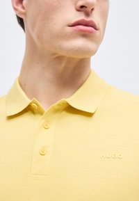 Jeune homme portant un polo jaune avec un col et trois boutons, arborant un petit logo "HUGO" sur la poitrine.
