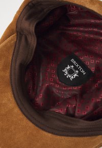 Rudono aksominio skrybėlės vidus su bordo pamušalu, turinčiu subtilų kryžiaus raštą, ir juodu �žymeniu, ant kurio nurodytas "Brixton" logotipas.