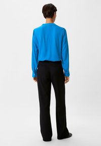 Camicetta blu a maniche lunghe con texture liscia e colletto, abbinata a pantaloni neri a gamba larga. La camicetta presenta polsini con bottoni.