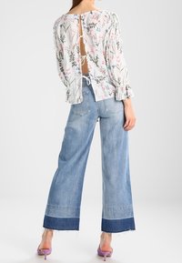 Blusa bianca trasparente a stampa floreale con chiusura sul retro; abbinata a jeans azzurri a gamba larga con orlo grezzo e dettagli in denim scuro.