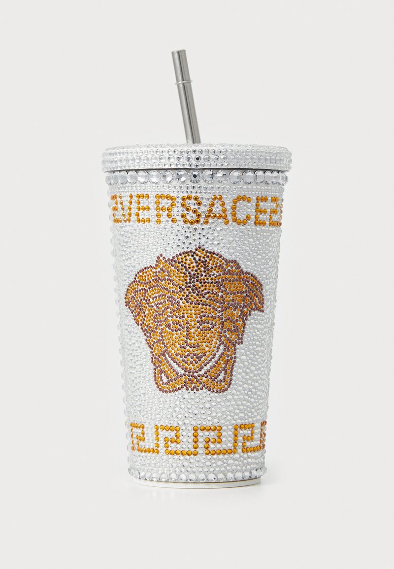 Cup argintiu decorat cu cristale, cu un logo de Medusa aurit, având un pai metalic și un model decorativ la bază.