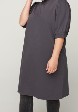 Kobieta w luźnej, ciemnoszarej sukience do kolan z bufkami na półrękawach, nosząca spodnie dżinsowe w ciemnym kolorze, stojąca na jednolitym tle.