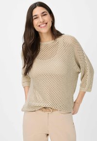 Beige gebreid trui met een open gatenpatroon, ruime halslijn en driekwartmouwen, gestyled met beige broeken en een riem.