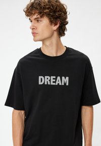 Mustapaita, jossa on valkoisella pistekuvalla varustettu "DREAM"-teksti etupuolella. Rentouttava malli, jossa on pyöreä kaula-aukko ja lyhyet hihat.