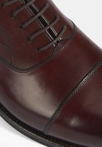 Chaussure habillée en cuir bordeaux avec une texture lisse, un embout arrondi et des coutures décoratives. Présente des lacets et une silhouette élégante, adaptée à une tenue formelle.