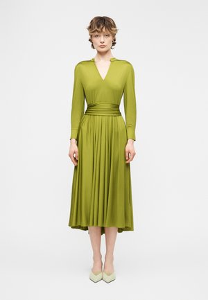 Vrouw in een olijfgroene midi-jurk met lange mouwen, een V-hals en een getailleerde taille, gecombineerd met lichtgekleurde puntige schoenen, staand tegen een witte achtergrond.