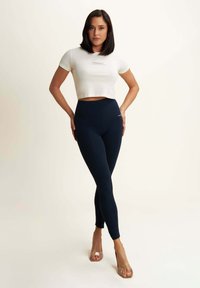 Marineblaue hochgeschnittene Leggings aus elastischem Material, kombiniert mit einem weißen, kurzen T-Shirt mit einem Logo. Transparente High-Heels.