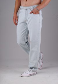 Mann mit hellblauen Jeans und weißen Sneakers, der mit einer Hand in der Tasche vor einem schlichten grauen Hintergrund steht.