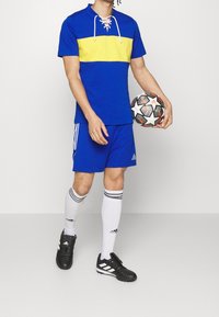 Modrý a žlutý sportovní tričko s krátkým rukávem a zavazováním u krku, doplněné modrými šortkami, černými kopačkami a bílými ponožkami s černými pruhy.