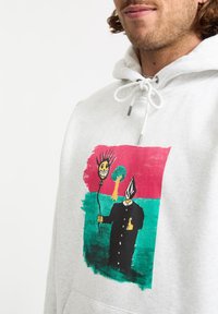 Haut à capuche gris avec un imprimé graphique coloré représentant une silhouette en noir tenant un ballon au visage souriant, le tout sur un fond multicolore.