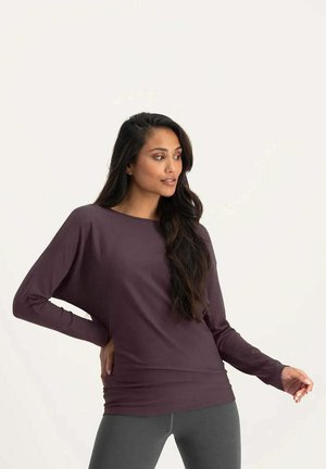 Frau mit langen dunklen Haaren, die ein locker sitzendes violettes Langarmshirt und graue Leggings trägt, posiert mit einer Hand in der Hüfte und schaut seitlich.